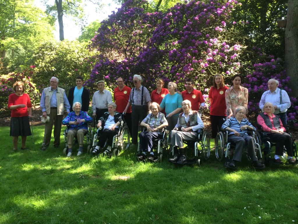 Gruppenfoto von Senioren, Pflegekräften und Angehörigen des Pflegedienstes Imeraj bei einem Ausflug in den Rhododendron Park Bremen. Die Gruppe sitzt und steht vor blühenden Rhododendronsträuchern im Sonnenschein.