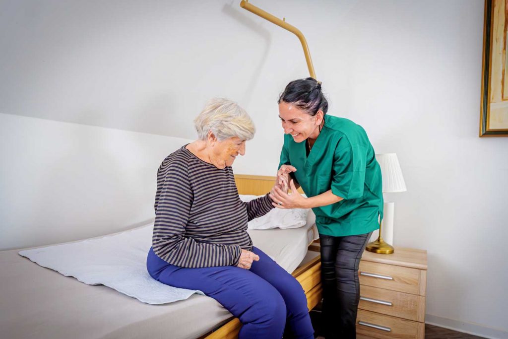 Eine Pflegefachkraft hilft einer Seniorin beim Aufstehen aus dem Bett in einem freundlich eingerichteten Zimmer.